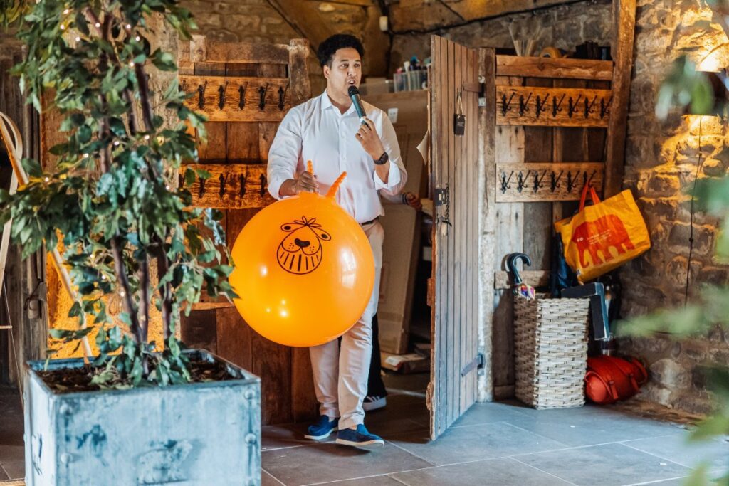 A man delivers a speech outdoors, holding a microphone and an orange balloon, surrounded by rustic wooden crates, potted plants, and festive decorations in a warmly lit setting at Tithe Barn, Skipton