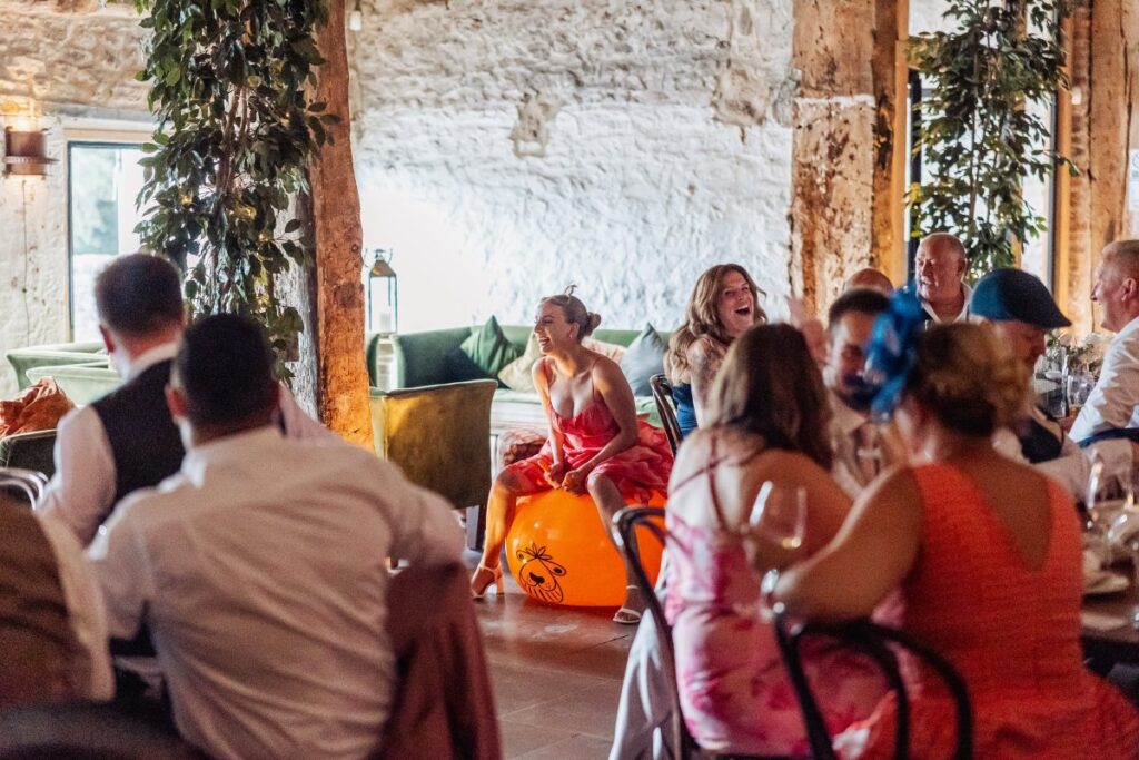 Guests gather for a lively wedding reception, sipping champagne and clapping as a bride in a flowing white gown sings along to the music, surrounded by bridesmaids in vibrant red at Tithe Barn, Skipton