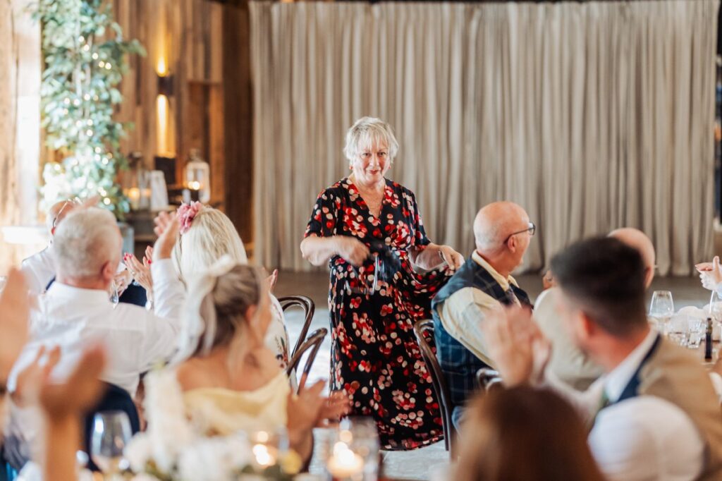 A guest delivers a heartfelt speech during wedding speeches, surrounded by clapping guests in a warmly lit room with elegant table settings and soft curtains at Tithe Barn, Skipton