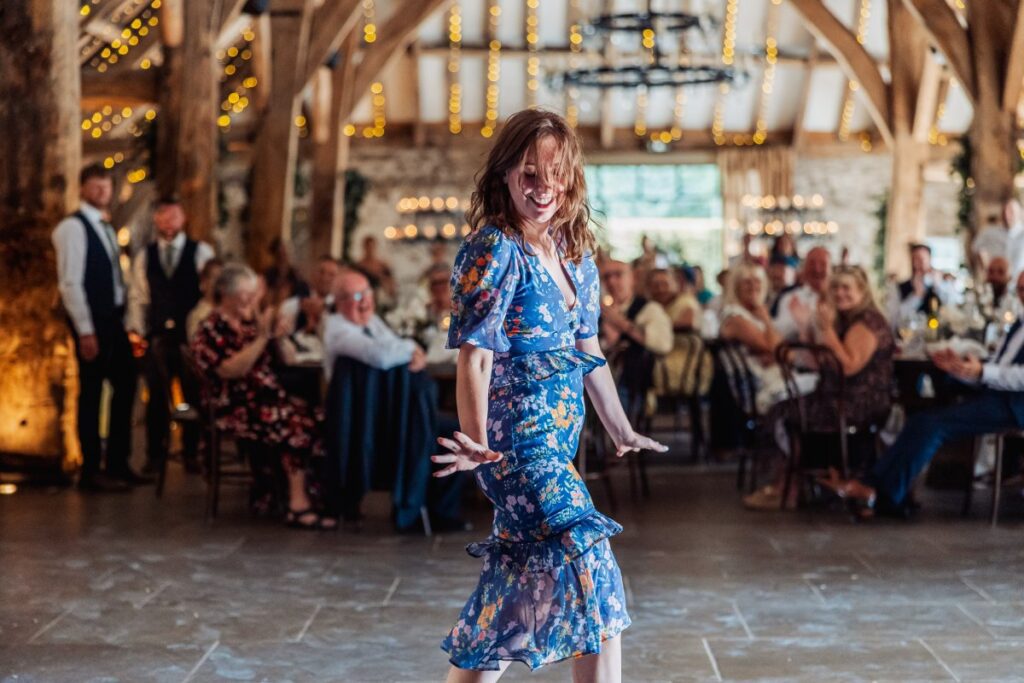 The bride dances joyfully in a blue floral gown as guests in formal attire cheer and applaud in a warmly lit wooden hall adorned with string lights at Tithe Barn, Skipton