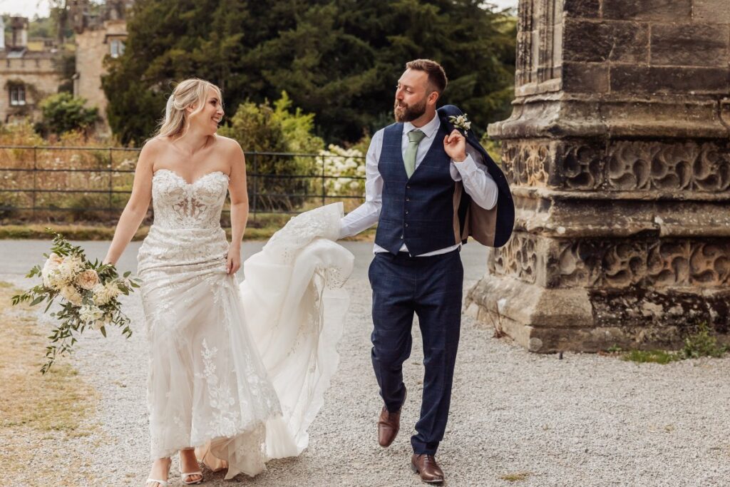 The bride, in a strapless lace gown and holding a cream bouquet, walks hand-in-hand with the groom in a navy suit and green tie, their joyful stride captured on a cobblestone path at Tithe Barn, Skipton