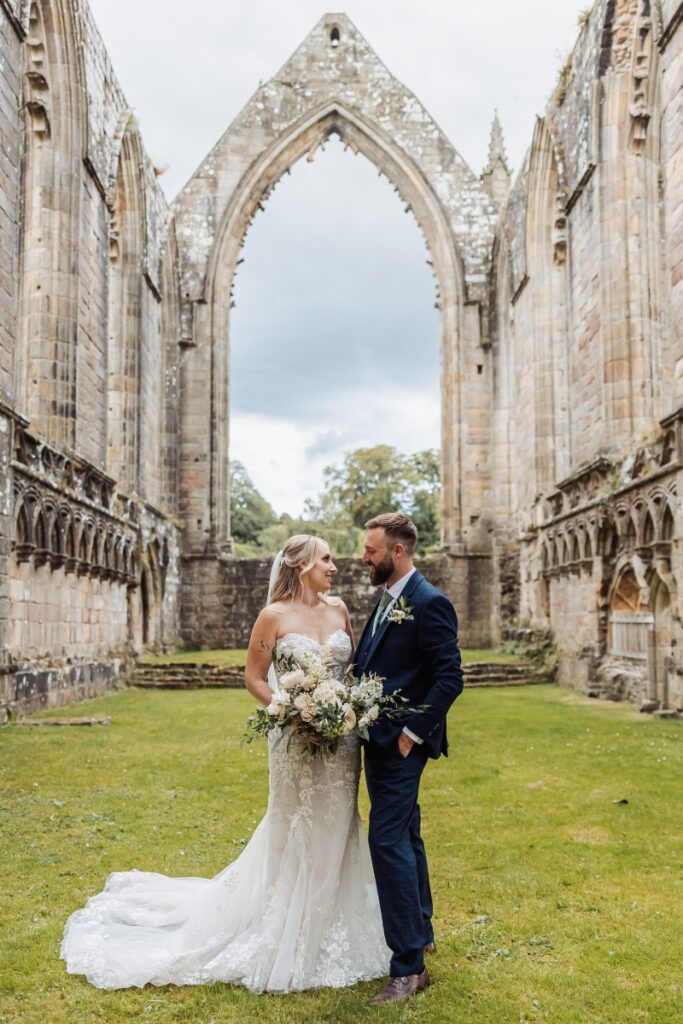 In a serene stone archway, the bride and groom share a tender moment, her white gown flowing gracefully as he holds her bouquet of cream and green blooms, the scene bathed in soft at Tithe Barn, Skipton