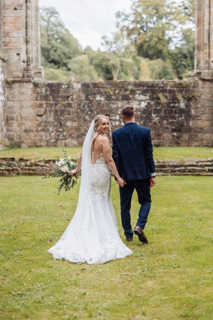 The bride and groom walk hand-in-hand across a lush green lawn, framed by ancient stone ruins and a backdrop of leafy trees, as they share a quiet moment after their ceremony at Tithe Barn, Skipton