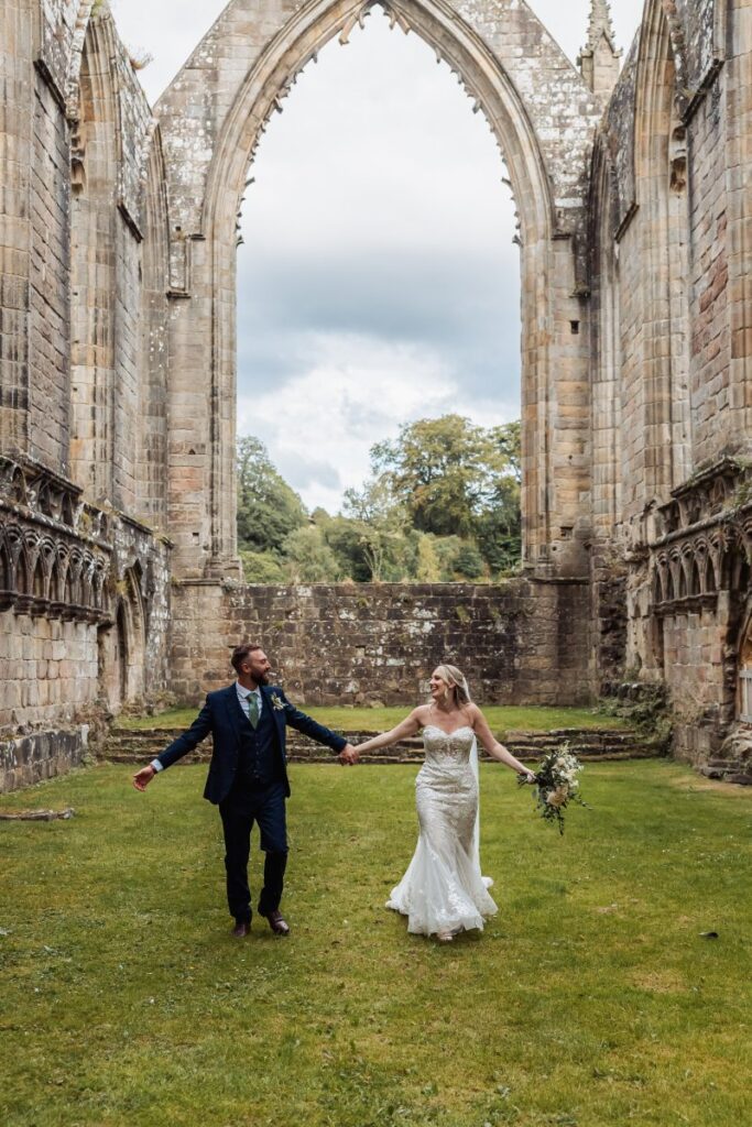 The bride and groom share a joyful first dance under a grand stone archway, their hands raised in celebration as they twirl on a lush green lawn, framed by ancient stone walls at Tithe Barn, Skipton
