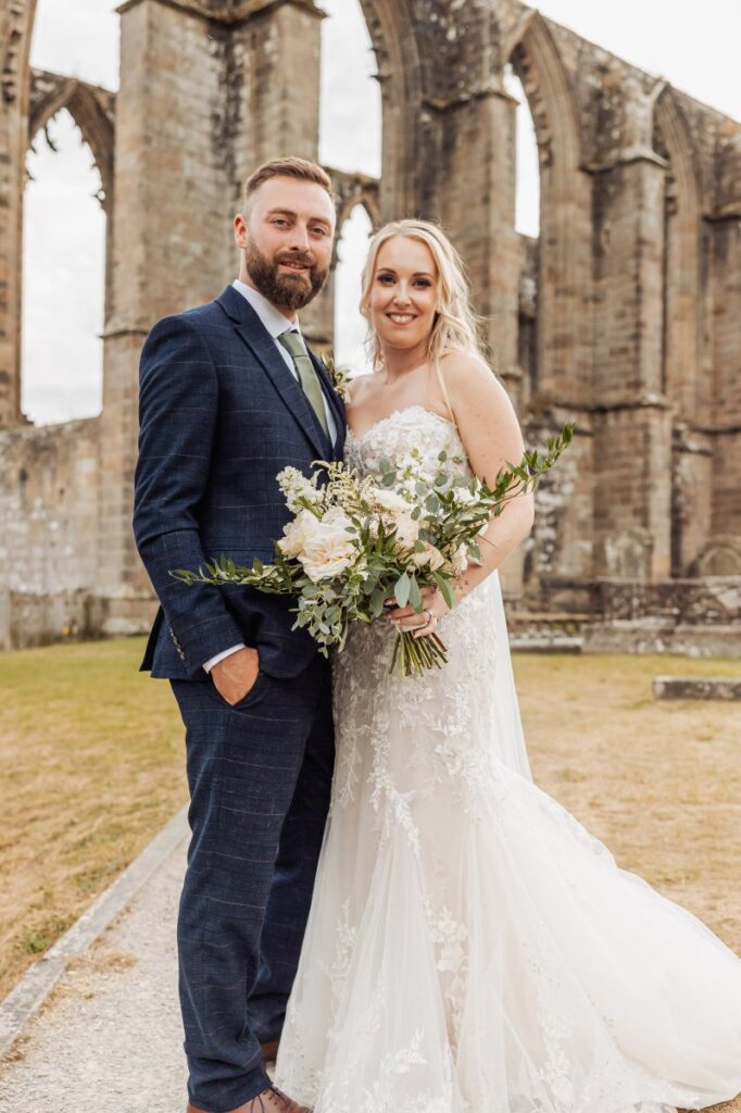 The bride, in a flowing white gown, holds a bouquet of cream and greenery as she and the groom, in a navy suit, stand arm in arm against the backdrop of a grand stone cathedral at Tithe Barn, Skipton