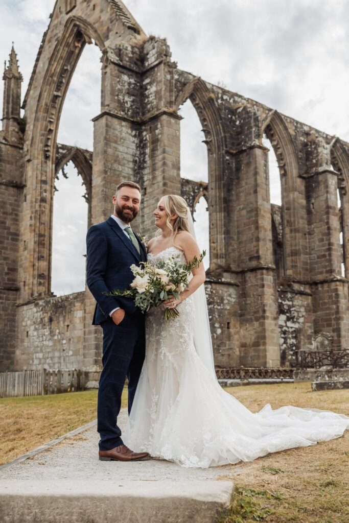 The bride and groom stand in front of a grand stone archway, the bride in a flowing white gown holding a bouquet of cream and green flowers, while the groom wears a navy suit and brown at Tithe Barn, Skipton