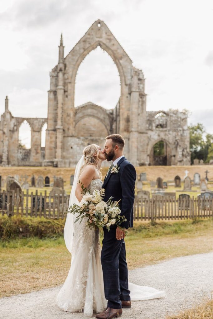 The bride, in a flowing lace gown, and groom, in a navy suit, share a tender kiss in front of a weathered stone arch, with a bouquet of cream blooms in her hands and a fence-lined path at Tithe Barn, Skipton