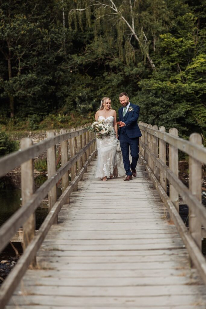 As they walk hand in hand across a rustic wooden bridge, the bride in a flowing lace gown and the groom in a navy suit stand out against the lush green backdrop of tall evergreen trees at Tithe Barn, Skipton
