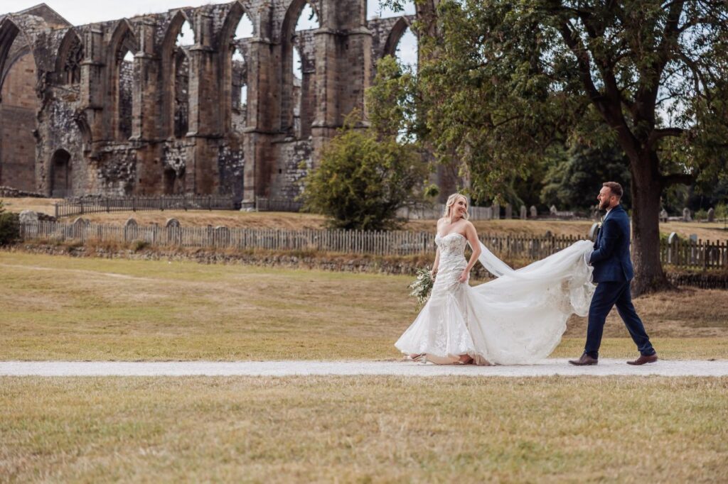 The bride and groom walk hand-in-hand across a grassy path, the bride's flowing white gown trailing behind as they embrace the serene setting of stone ruins and leafy trees at Tithe Barn, Skipton