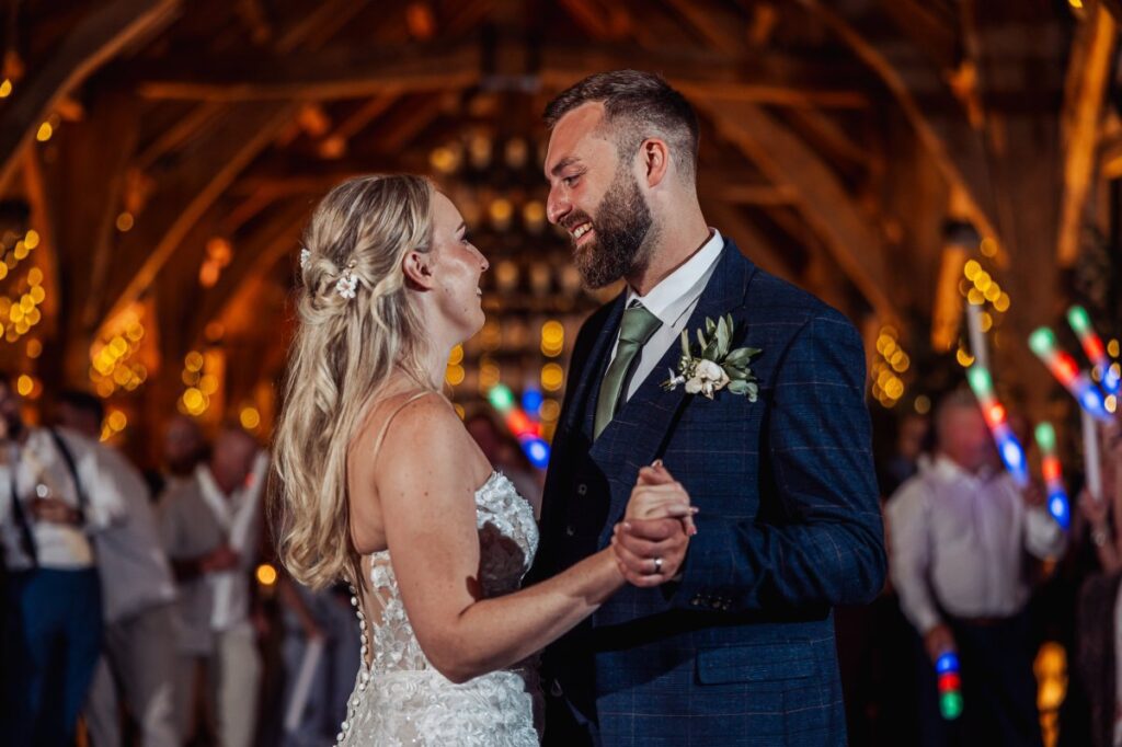 The bride and groom share their first dance in a warmly lit wooden hall, surrounded by guests holding colorful light sticks and a backdrop of twinkling fairy lights at Tithe Barn, Skipton