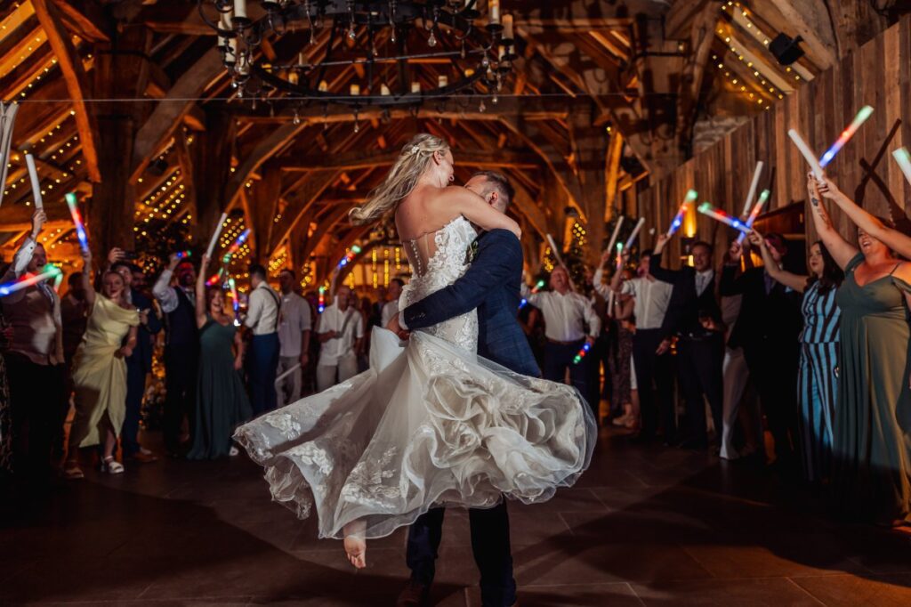Guests wave colorful glow sticks as the bride and groom share their first dance in a warmly lit, wood-paneled hall at Tithe Barn, Skipton