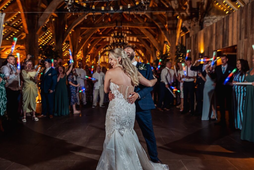 Guests wave colorful light sticks as the bride and groom share their first dance in a warmly lit wooden hall, surrounded by friends and family dressed in formal attire at Tithe Barn, Skipton