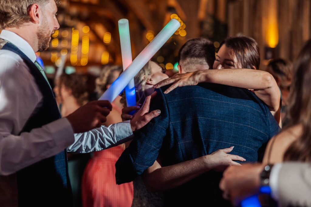 Amidst a lively dance floor illuminated by glowing LED sticks, the bride and groom embrace tightly as guests surround them, celebrating their first dance in a warmly lit wooden hall at Tithe Barn, Skipton