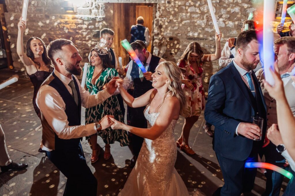 Confetti rains down as the bride in a shimmering gown and groom in a vest and bow tie share their first dance under warm, festive lights, surrounded by guests waving glow sticks at Tithe Barn, Skipton
