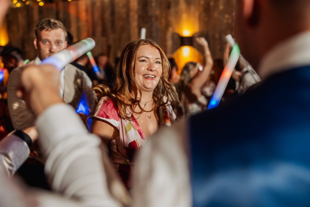 Guests dance joyfully during the first dance, waving glowing light sticks under warm, golden lighting in a wood-paneled hall at Tithe Barn, Skipton