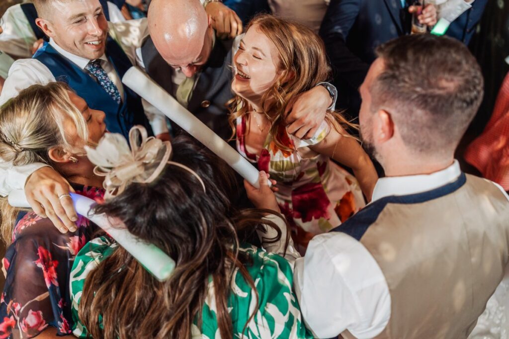 Guests gather around the bride and groom as they cut the cake, their hands reaching out to share in the moment, while a child in a patterned dress smiles brightly at Tithe Barn, Skipton