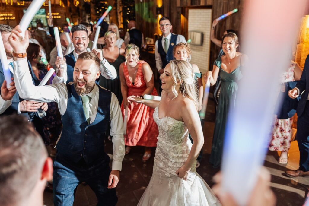 Amidst a vibrant first dance, the bride in a shimmering lace gown and groom in a navy suit celebrate with guests waving colorful glow sticks, their joy mirrored by the festive at Tithe Barn, Skipton