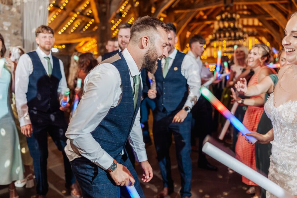 Guests dance joyfully under warm, golden lights, wielding colorful glow sticks as the bride and groom lead the celebration in a rustic wooden hall at Tithe Barn, Skipton