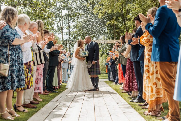 Guests applaud as the bride in a flowing white gown and the groom in a navy suit and kilt share a tender kiss under a canopy of dappled sunlight in a serene garden setting at Alexander Weddings & Events, Northallerton