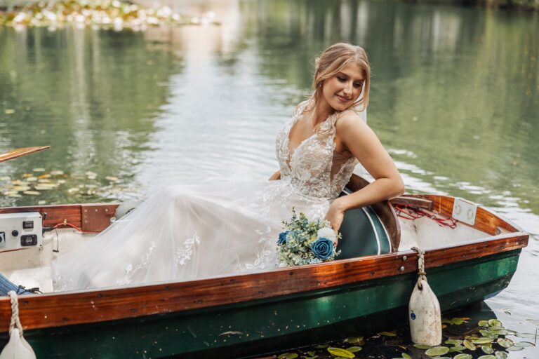 The bride sits gracefully in a wooden rowboat on a tranquil lily pond, her lace gown flowing around her as she holds a bouquet of blue and white flowers at Alexander Weddings & Events, Northallerton