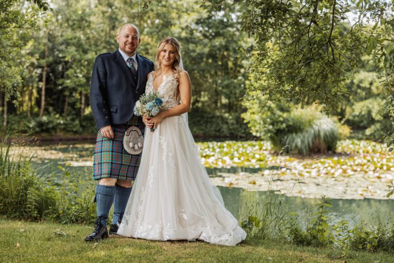The bride, dressed in a flowing white gown, stands beside her groom in a navy suit and tartan kilt, their hands clasped as they pose in a sun-dappled garden filled with vibrant yellow at Alexander Weddings & Events, Northallerton