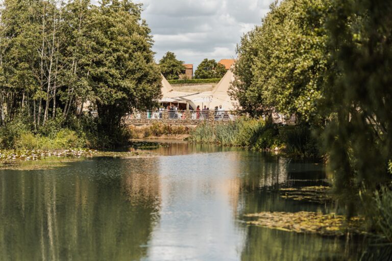 The bride and groom share their first dance under a canopy of trees, their silhouettes reflected in the tranquil water of a lily pond as golden hour light bathes the scene at Alexander Weddings & Events, Northallerton