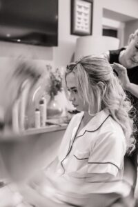 A woman in a white satin pyjama top is seated, looking down, as another person styles her hair in a well-lit room