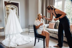 A bride in a white wedding dress is getting ready, seated in a chair, while a woman applies makeup to her face
