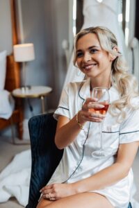 A woman in a white satin nightshirt holds a glass of pink champagne, seated on a dark chair in a warmly lit hotel room