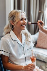 A woman in a white satin pyjama top smiles as she holds a glass of rose wine while having her makeup applied at Woodlands Hotel, Leeds