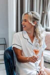 A woman in a white satin pyjama top sits on a blue velvet chair, holding a glass of rose wine and smiling