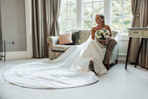 A bride in a white wedding dress sits on a floral-patterned armchair, holding a bouquet, smiling and looking to the side