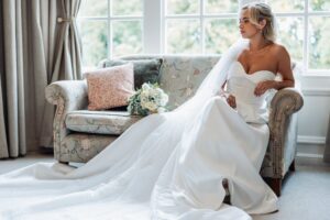 A bride in a strapless white wedding gown with a long veil is seated on a floral-patterned armchair, holding the train of her dress at Woodlands Hotel, Leeds
