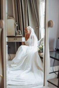A bride in a white wedding dress with a long veil sits on a couch, holding a bouquet of flowers, in a well-lit room with curtains and a dressing table