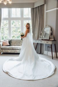 A bride in a white wedding dress stands in a room with a large window, holding a bouquet of flowers at Woodlands Hotel, Leeds