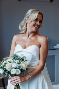 A woman in a strapless white wedding dress holds a bouquet of white flowers and greenery, smiling warmly