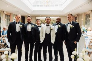 A group of men in formal attire, including tuxedos and a white suit, are standing together in a well-lit indoor wedding venue