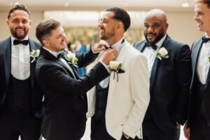 A man in a white suit has his bow tie adjusted by another man in a black tuxedo, while others in formal attire look on with smiles at Woodlands Hotel, Leeds