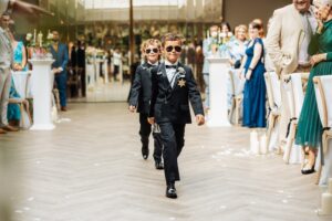 Two young boys in matching navy suits and sunglasses walk down a wooden floor in a well-lit indoor venue, surrounded by seated guests in formal attire