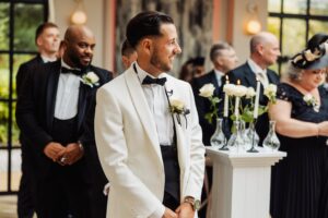 A man in a white tuxedo with a black bow tie stands in front of a group of seated guests in formal attire at Woodlands Hotel, Leeds
