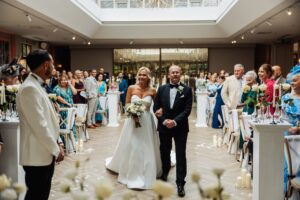 A bride in a white gown and bouquet walks down the aisle with an older man in a dark suit