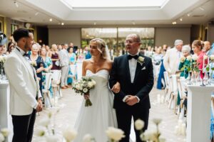 A bride in a strapless white gown walks down an aisle with an older man in a dark suit at Woodlands Hotel, Leeds