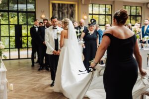 A couple in wedding attire stands facing each other, holding hands, with an officiant and guests seated in the background at Woodlands Hotel, Leeds