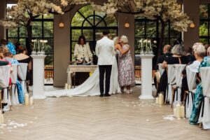A couple stands facing an officiant at a wedding ceremony, with guests seated on either side at Woodlands Hotel, Leeds