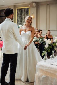 A couple stands facing each other, holding hands, in a warmly lit room during a wedding ceremony
