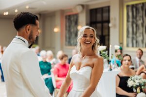 A couple in wedding attire stands facing each other during a ceremony, with guests seated in rows and white flowers and candles at Woodlands Hotel, Leeds