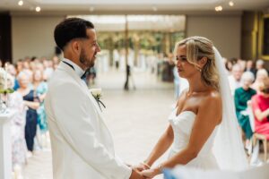 A couple in wedding attire stands facing each other, holding hands, with guests seated in rows behind them