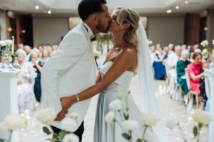 A couple in wedding attire share a kiss during a ceremony, with guests seated in rows and white flowers and candles on the tables