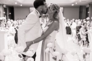 A couple in wedding attire share a kiss during a ceremony, with guests seated in rows in the background at Woodlands Hotel, Leeds