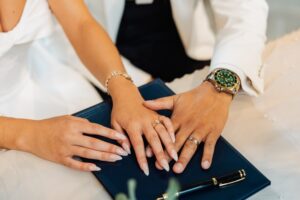 Two people in wedding clothes hold hands on a dark blue folder with a pen, wearing rings and a wristwatch at Woodlands Hotel, Leeds